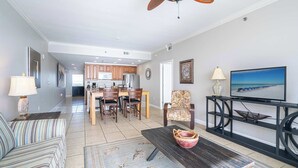 Amplia sala de estar con sofá, mesa de centro y mueble para televisión. También se aprecia un comedor con sillas y una cocina al fondo.