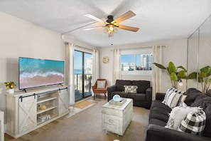 A living room with a television, a ceiling fan, a sofa, a coffee table, and a view of the ocean through a sliding door.