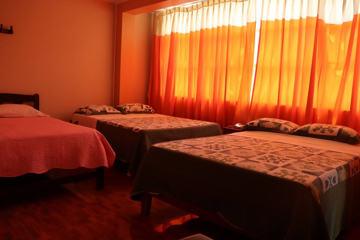 A hotel room with two single beds, each with a patterned bedspread and pillows. The room has wooden flooring and a window with orange curtains.