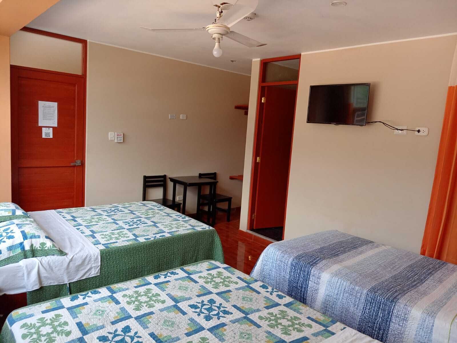 A room with two beds, a ceiling fan, a TV mounted on the wall, and a small table with chairs.