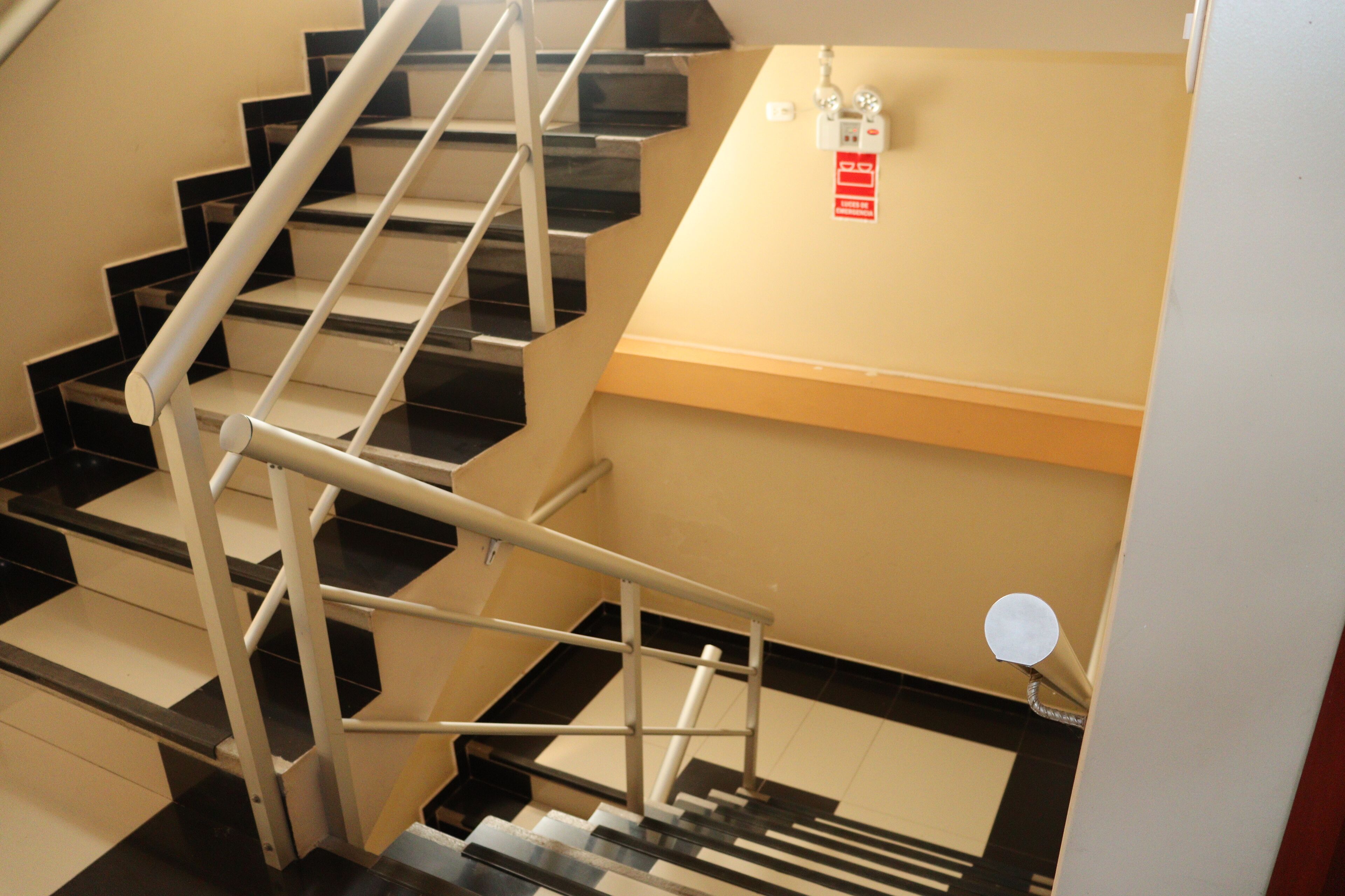 A set of black and white stairs with a metal handrail, a beige wall, and a fire alarm on the landing.
