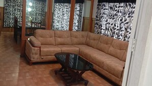 Ένα σαλόνι με έναν καφέ γωνιακό καναπέ, ένα τραπεζάκι σαλονιού από γυαλί και κουρτίνες με σχέδια.