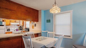 Una cocina con armarios blancos, una mesa de comedor blanca y sillas. Hay una ventana con persianas y una lámpara colgante.