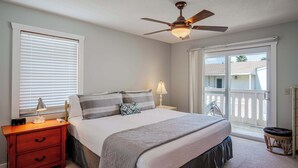 Un dormitorio con cama, mesita de noche, lámpara, ventilador de techo y vistas a través de un ventanal corredero.