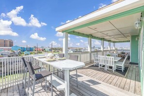 A covered outdoor deck with a white table and chairs, a view of the ocean, and a green building in the background.