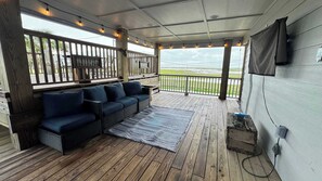 A covered outdoor seating area with a sofa and a TV, wooden deck flooring, and a view of the landscape.