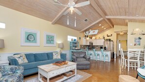 A spacious living room with a high ceiling, a blue sofa, a coffee table, and a kitchen area in the background. - Delightful Stays 2 Bdr | Pool | | Sleeps 8 (Port Aransas)