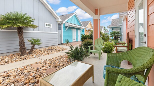 A covered patio with wicker furniture, a palm tree, and a small garden area with rocks and plants. - Delightful Stays 2 Bdr | Pool | | Sleeps 8 (Port Aransas)