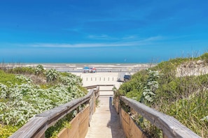 Ein Holzweg führt zu einem Strand mit freiem Blick auf das Meer und einigen Sonnenschirmen.