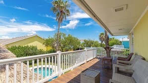 A wooden deck with a white railing, wicker furniture, and a view of a swimming pool and greenery. - Private Pool | Pets | Sleeps 7 | Long Term Stays (Port Aransas)