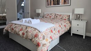 Een net opgemaakt bed met een bloemenprint deken, een nachtkastje met een lamp, en een schilderij aan de muur.