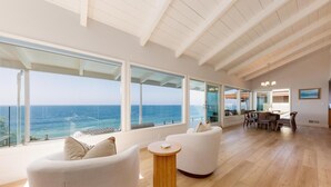 A spacious living area with large windows overlooking the ocean, a round wooden table, and a dining area with chairs. - Oceanfront Luxury with Pool/Spa, Beach Access (Encinitas)