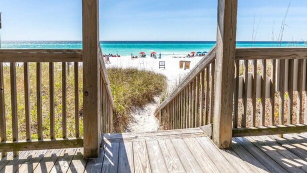 A wooden deck leading to a beach with clear turquoise water and a sandy shore.