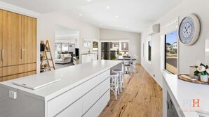 A modern kitchen with a large island, wooden cabinets, and a dining area with white chairs. - Coastal Family Retreat in CW (Cape Woolamai)