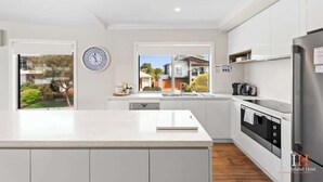 A modern kitchen with white cabinetry, a large island, a built-in oven, and a view of a residential area. - Coastal Family Retreat in CW (Cape Woolamai)