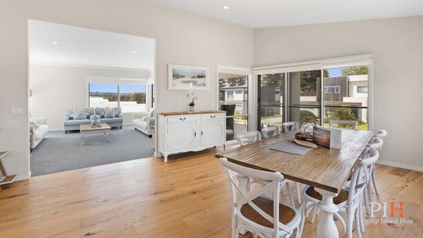A spacious living area with a wooden dining table, white chairs, a sofa, and a kitchen area with a white cabinet. - Coastal Family Retreat in CW (Cape Woolamai)