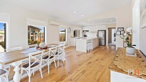 A spacious kitchen with white cabinetry, a central island, and a dining area with a wooden table and white chairs. - Coastal Family Retreat in CW (Cape Woolamai)