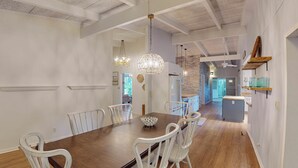 Un coin repas avec une table en bois et des chaises blanches, un luminaire suspendu et une cuisine en arrière-plan.