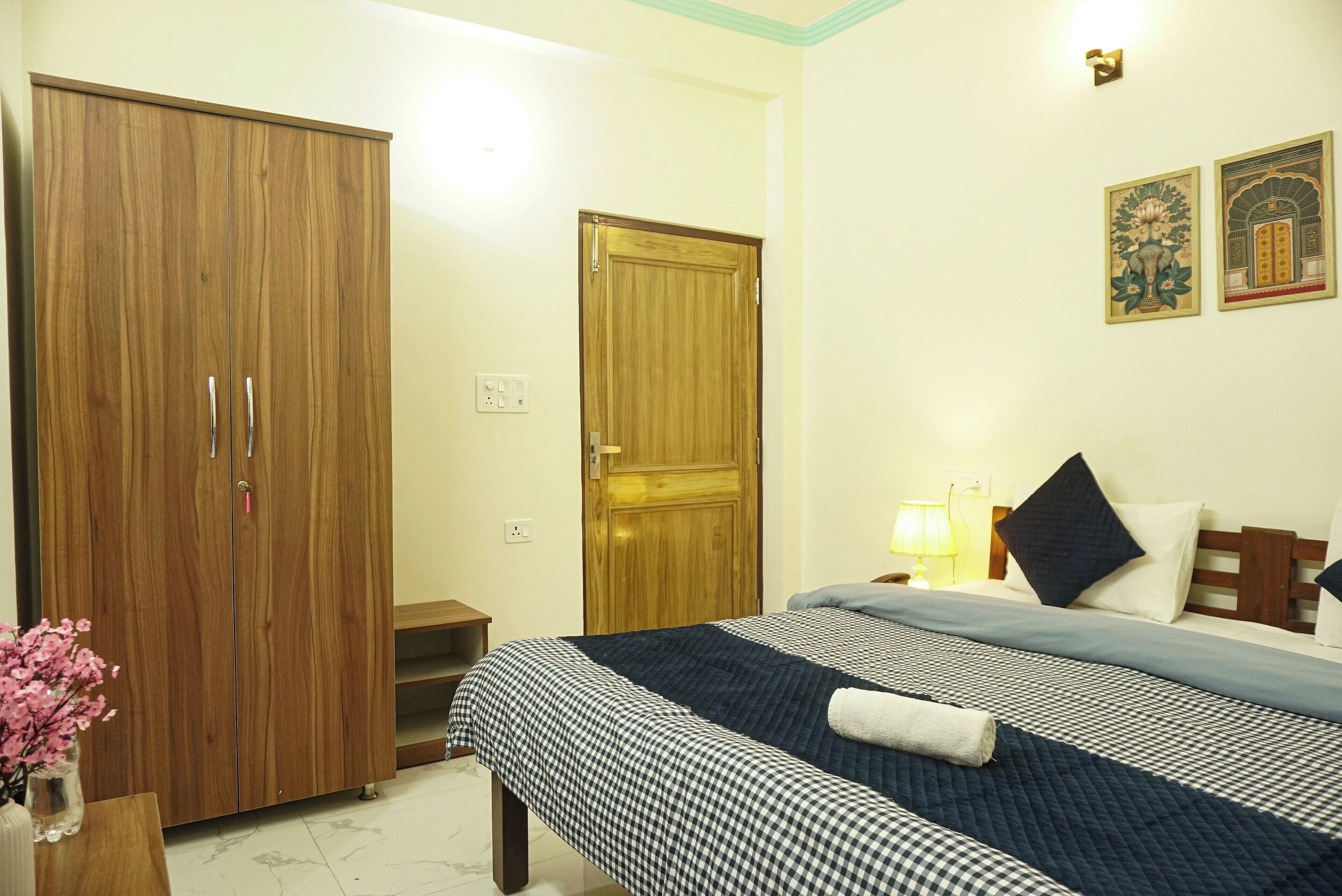 A hotel room with a large wardrobe, a bed with a checkered bedspread, a nightstand with a lamp, and two wall-mounted artworks.