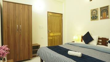 A hotel room with a large wardrobe, a bed with a checkered bedspread, a nightstand with a lamp, and two wall-mounted artworks.