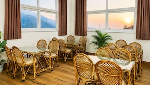 A dining area with wicker furniture, large windows with curtains, and a view of a sunset. - Anandmaya Retreat Tapovan Rishikesh (Rishikesh)