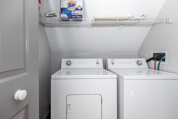 Une buanderie équipée de deux machines à laver à chargement frontal blanches, d’une étagère sur laquelle se trouve une boîte de lessive Tide, et d’une bouche d’aération au mur.