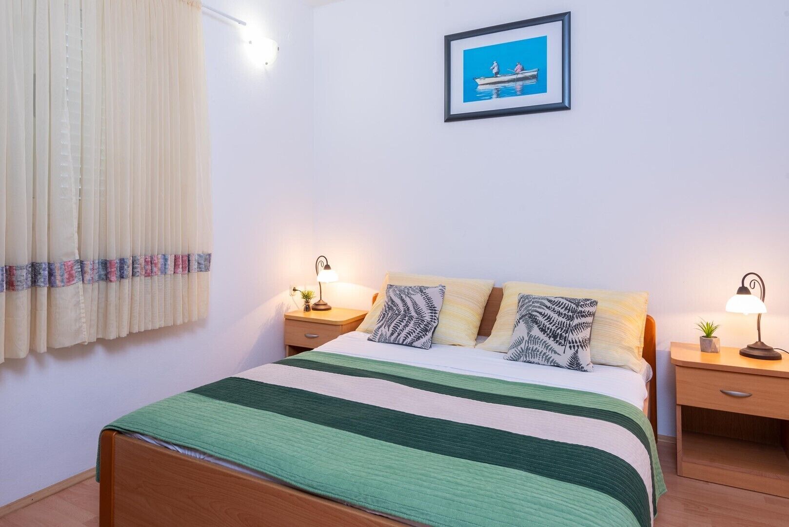 A neatly made bed with a striped bedspread, two bedside lamps, a framed picture on the wall, and a window with curtains.