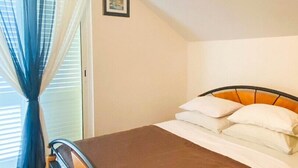 Un llit individual amb un estovat marró i coixins blancs en una habitació amb terra de fusta i una finestra amb cortines blaves.