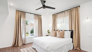 A bedroom with a large bed, beige curtains, a ceiling fan, and a window with a view of the outside.