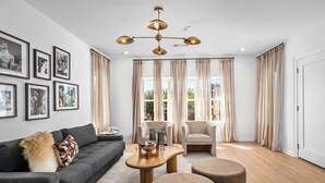 A modern living room with a grey sofa, two armchairs, a wooden coffee table, and round ottomans. There are framed pictures on the wall, a ceiling light fixture, and large windows with curtains.