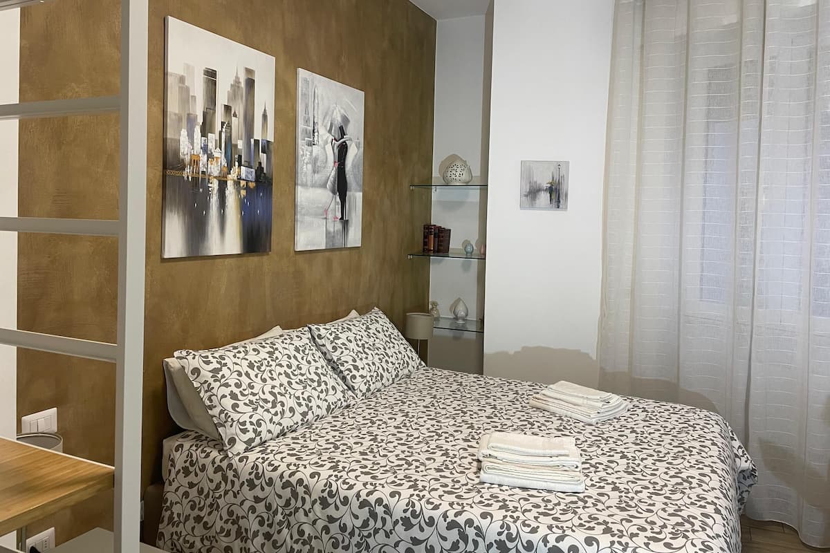 Quarto com cama, mesa de cabeceira, prateleira com objetos decorativos e duas pinturas penduradas na parede.