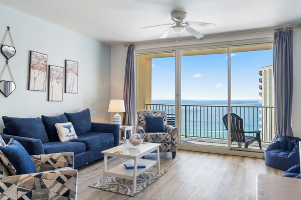 Sala de estar com sofá, poltrona e mesa, varanda com vista para o mar e ventilador de teto.