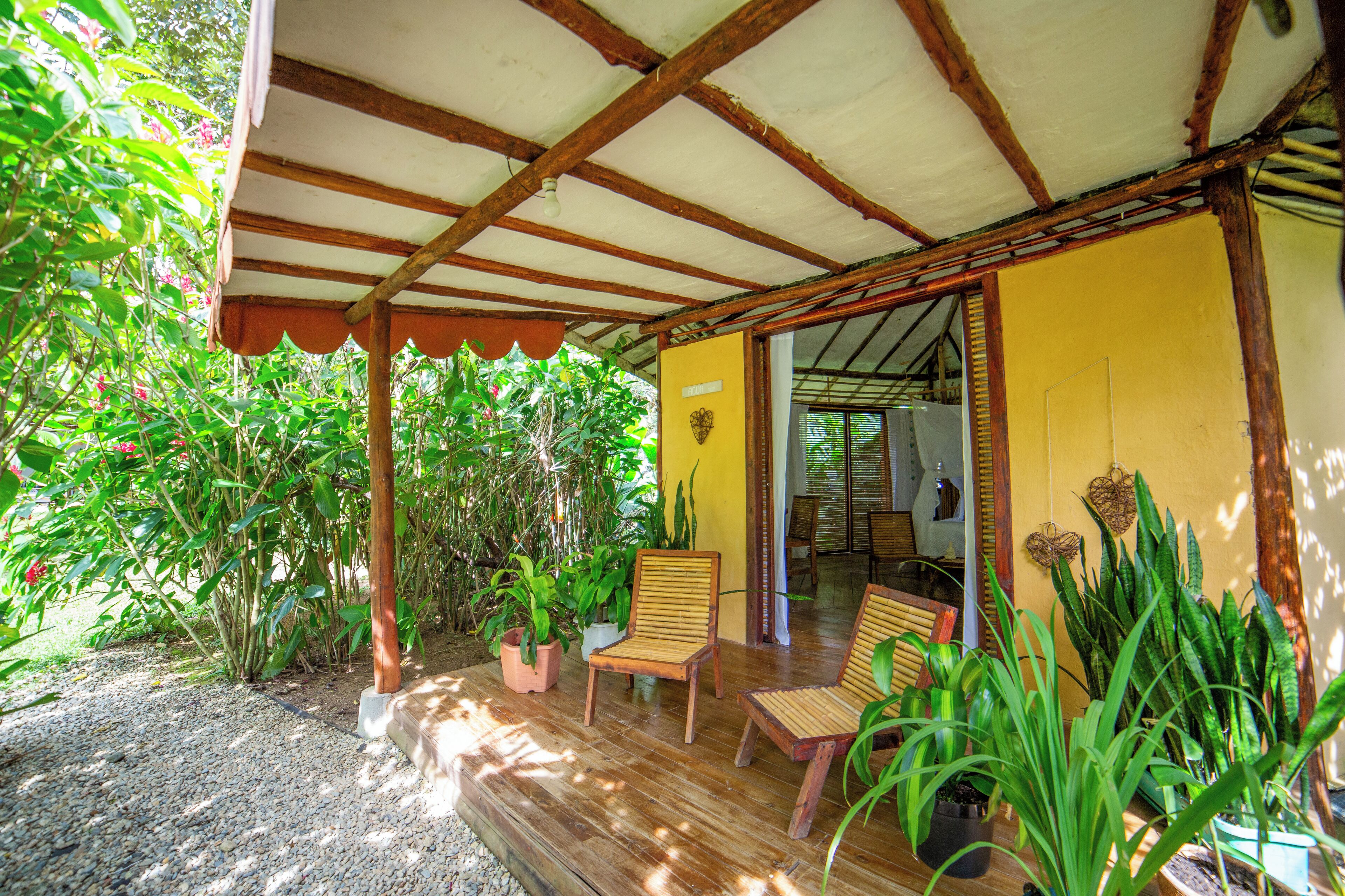 A covered outdoor seating area with wooden chairs and potted plants, a yellow wall, and a wooden ceiling structure.