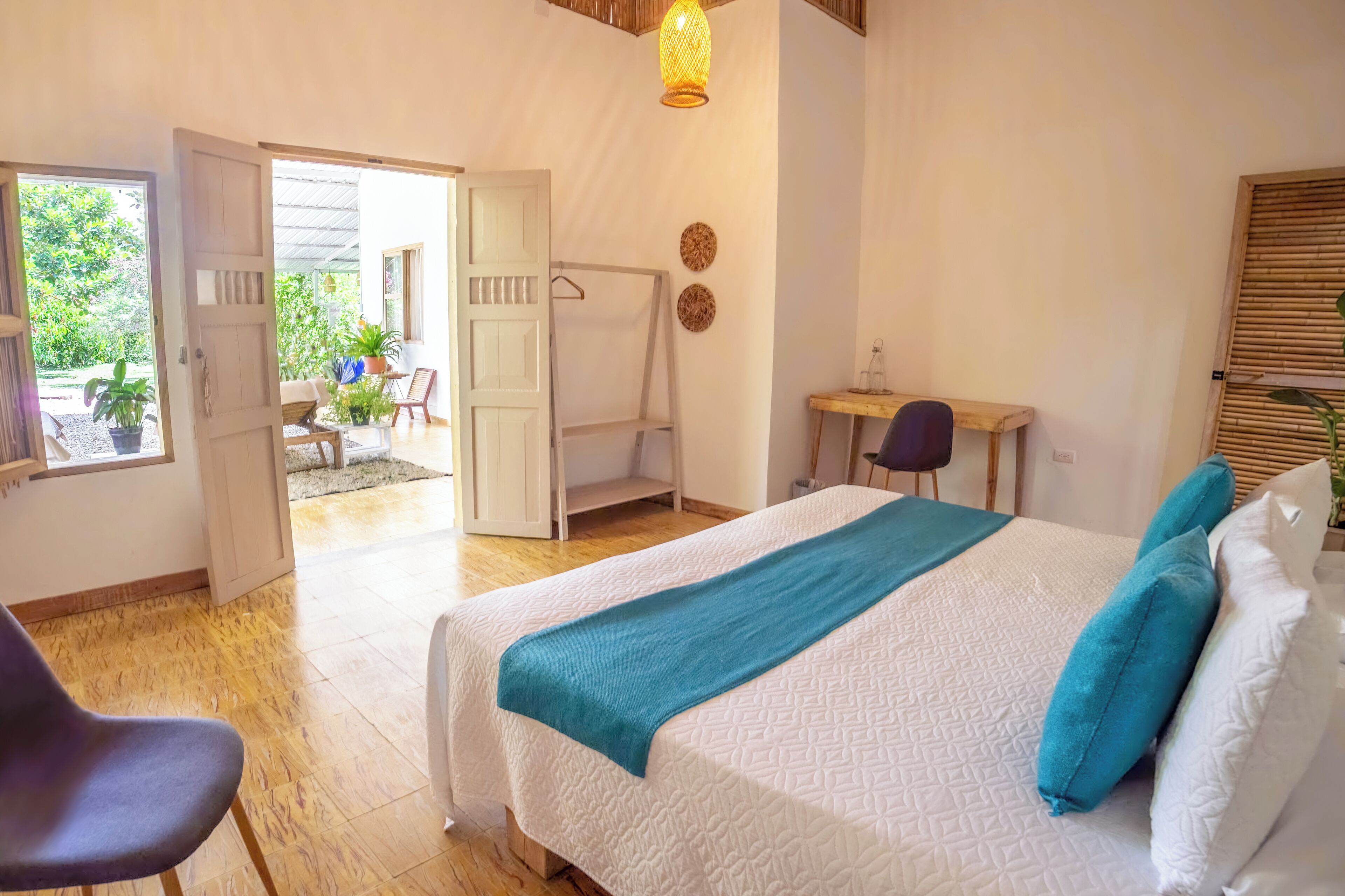 A bedroom with a bed, a desk, and a chair. There is a door leading to an outdoor area with plants.