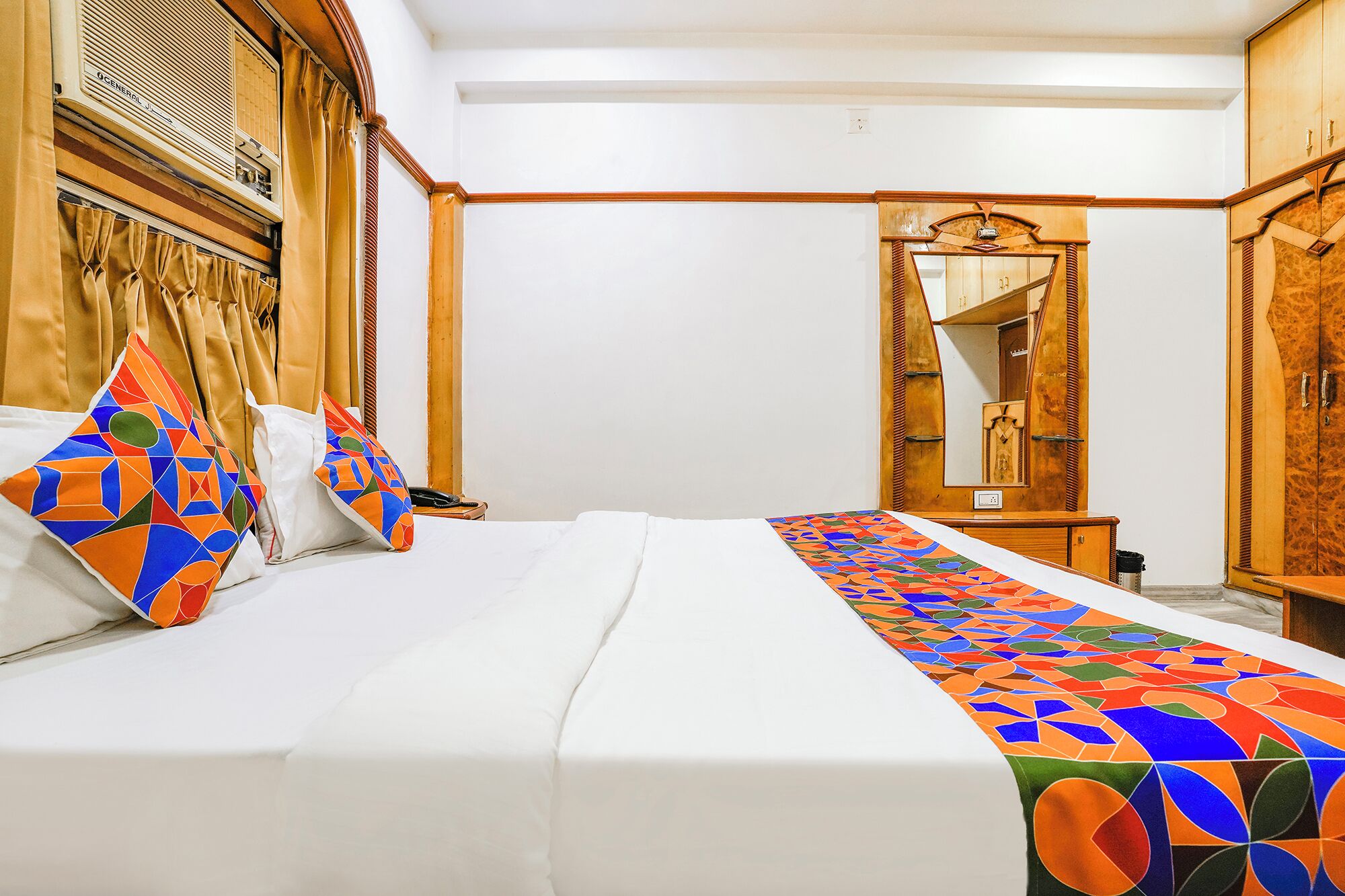 A hotel room with a bed, a wooden wardrobe, a mirror, and a patterned bed runner.