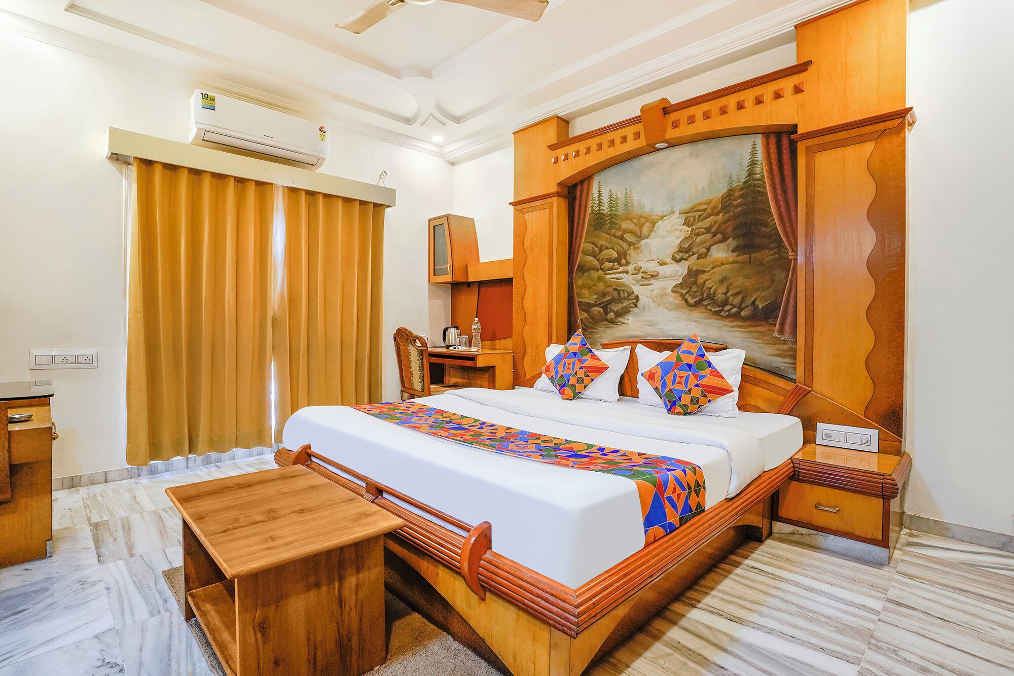 A hotel room with a large bed, a wooden headboard, a painting above the bed, a desk with a chair, and an air conditioning unit.