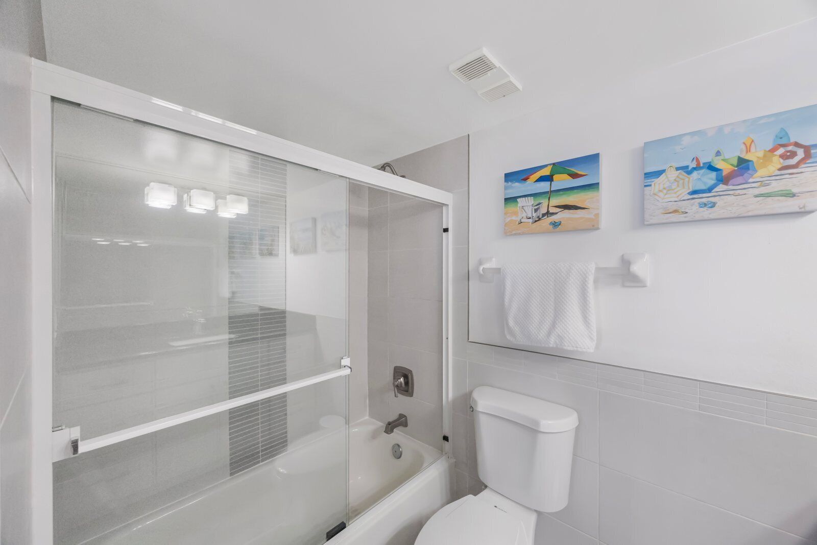 Un baño moderno con ducha envuelta en vidrio, bañera y retrete. Hay dos toalleros de pared y un cuadro de una playa.