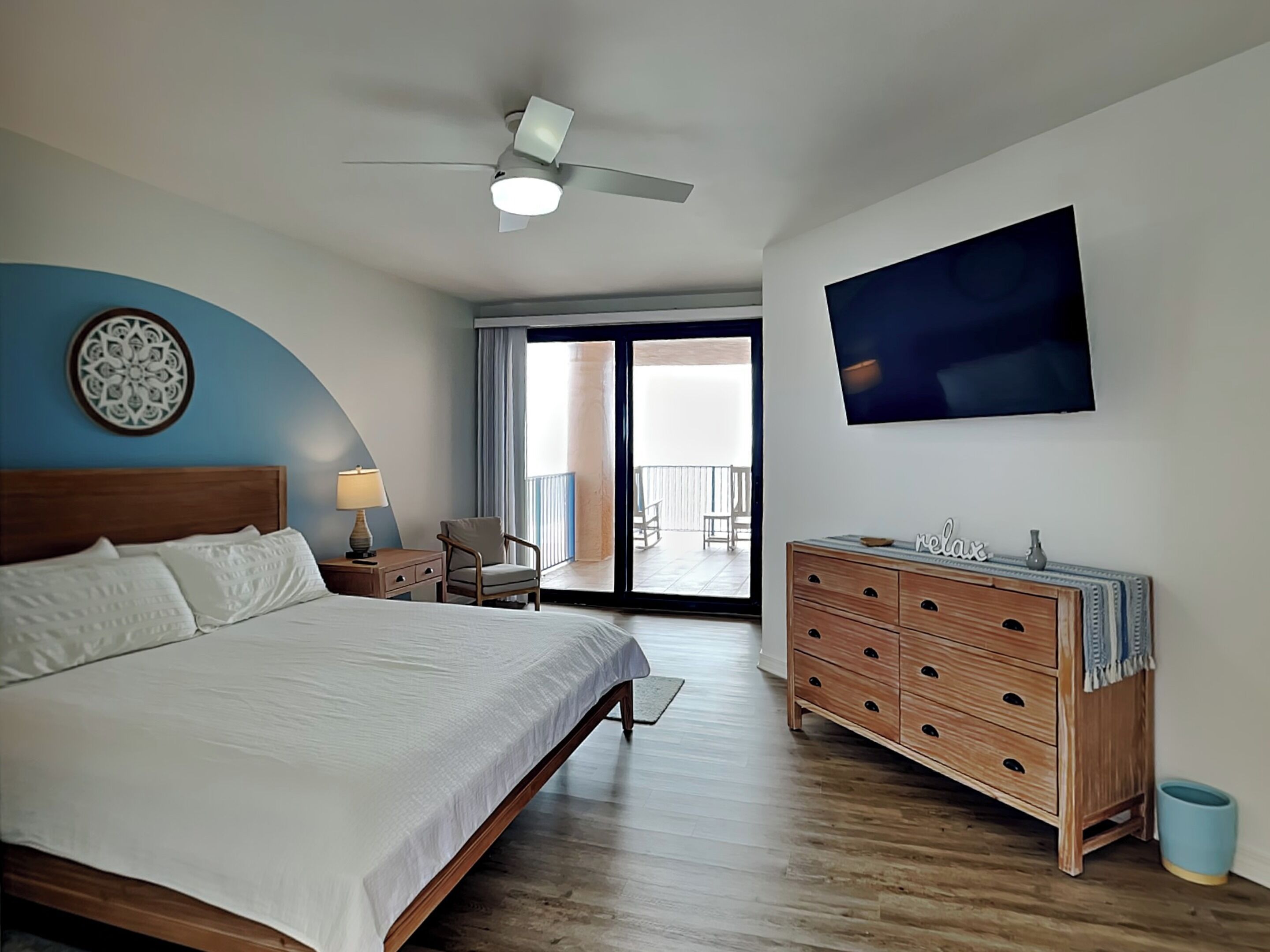 A bedroom with a bed, a dresser, a ceiling fan, a TV, a balcony with a view, and a decorative wall piece.