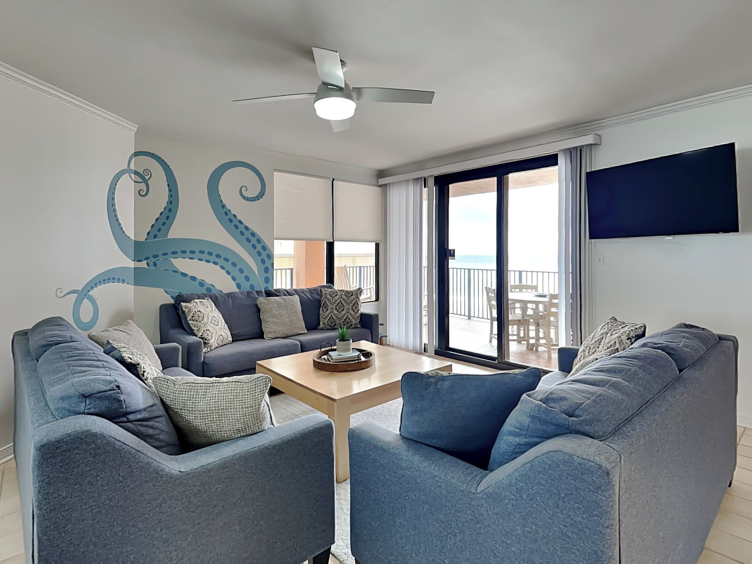 A modern living room with a ceiling fan, a large TV, and a view of the balcony.
