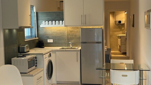 Une cuisine compacte équipée d’un lave-linge, d’un four à micro-ondes et d’un réfrigérateur. On y trouve une table en verre avec une chaise, ainsi qu’un petit coin salon avec un canapé.