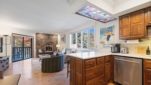 A modern kitchen with wooden cabinets, a stainless steel dishwasher, and a large island with drawers. There is a stone wall and a large window with a view of trees.