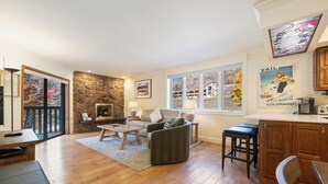 A spacious living room with a stone fireplace, a large window with a view of a building, and a kitchen area with wooden cabinets.