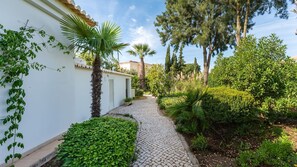 A paved pathway leading to a white building with a tiled roof, surrounded by lush greenery and trees.
