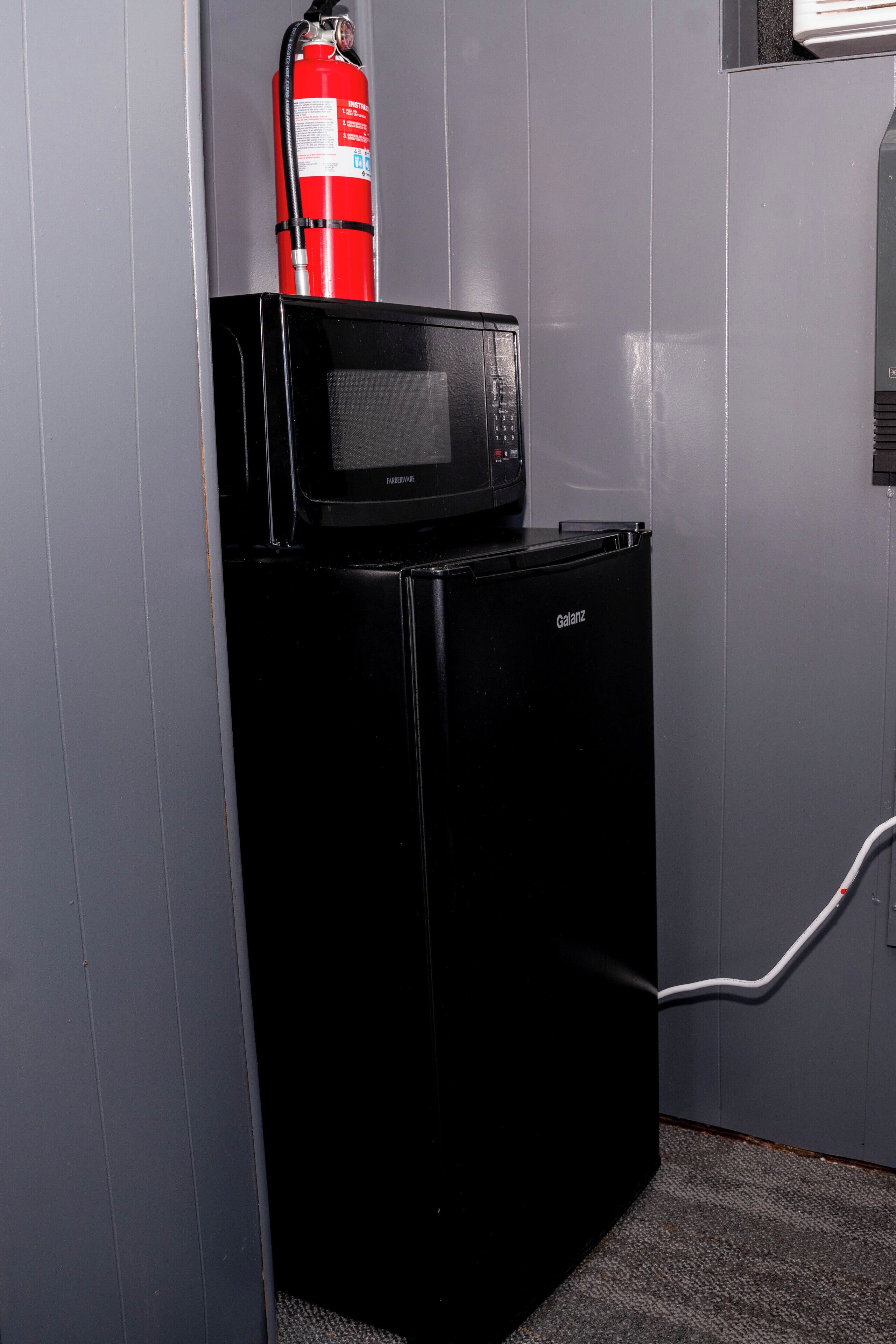 A black refrigerator with a microwave on top, a fire extinguisher mounted on the wall, and a white cable running along the wall.