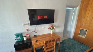 A room with a wooden desk, a television mounted on the wall displaying 'NETFLIX', a small vase with flowers, a remote control, and a coffee maker. - Maverick Motel and Glamp Ground (Raton)