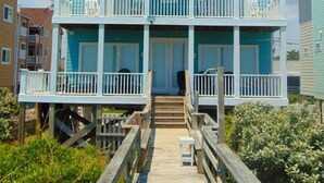 A two-story beachfront house with a wooden deck and a balcony.