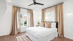 Una camera da letto con un letto, tende color beige, una ventola a soffitto e una finestra vista casa.