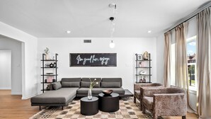 Un soggiorno moderno con un divano componibile, due poltrone, un tavolino da caffè e un tappeto decorato. Si vedono librerie con oggetti ornamentali e un lampadario a soffitto.