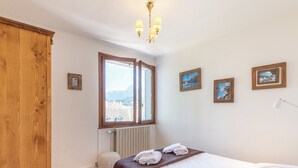 A bedroom with a wooden wardrobe, a large window with a view of buildings, a radiator, and a bed with towels.