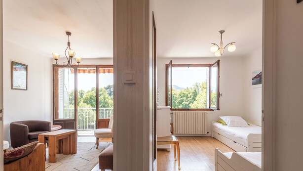 A room with a wooden floor, a white bed, a chair, a table, a radiator, and a window with a view of trees.